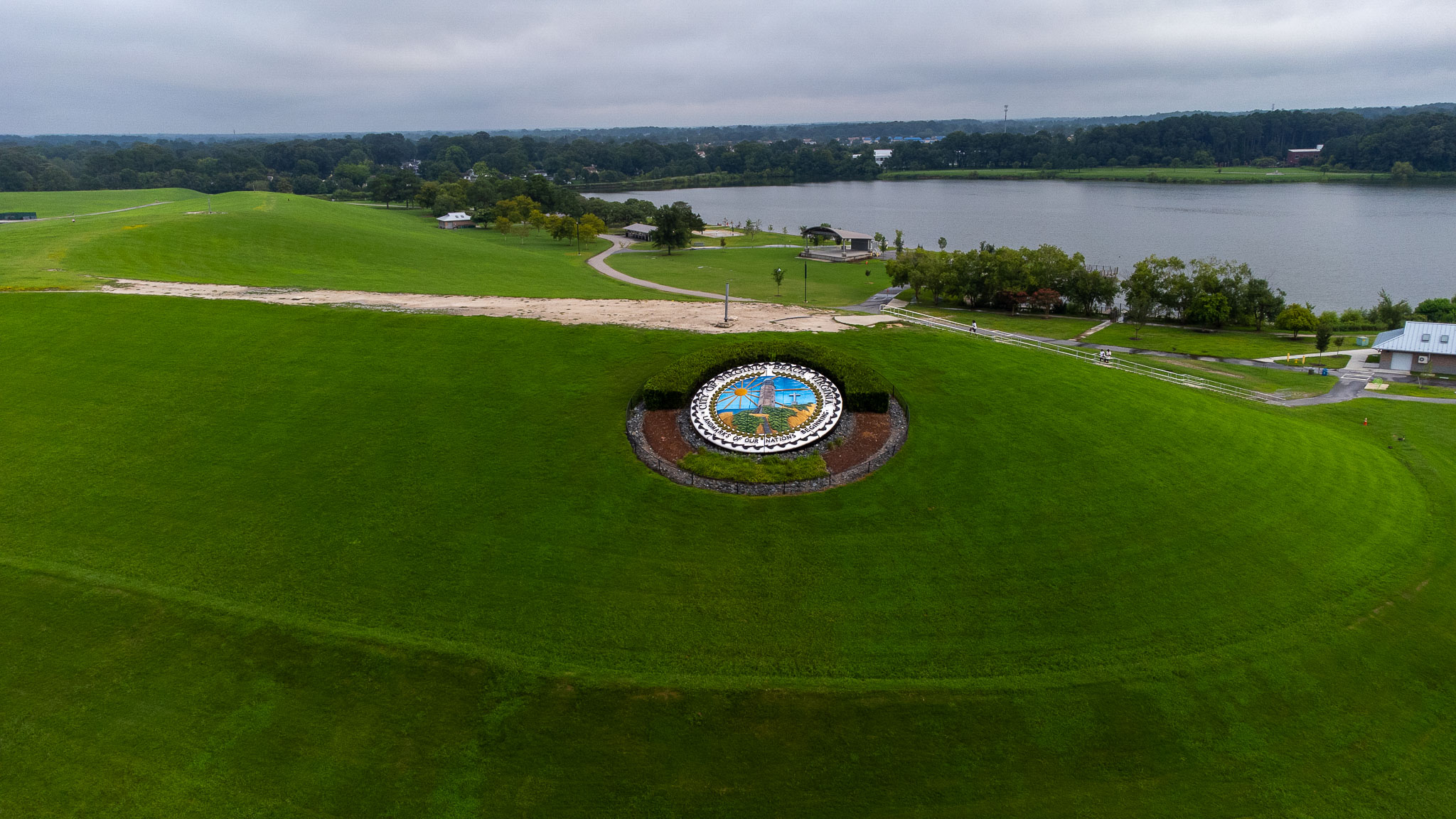 Aerial view of a colorful circular medallion embedded in the grassy embankment of a dam next to a lake under an overcast sky.